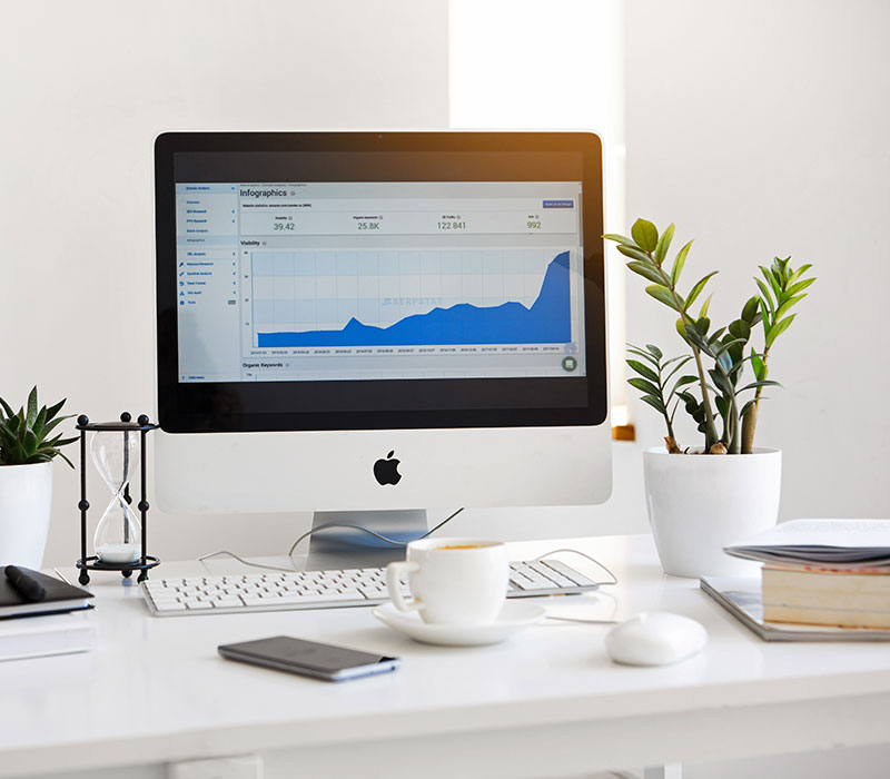 Desktop computer, flower pots, white cup of tea, a phone and a book on a table.